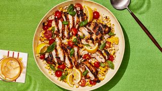 Sliced grilled chicken with corn and tomatoes over couscous with lemons in a big serving platter on a green tablecloth.